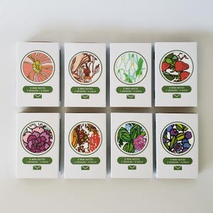 May include: Eight mini note cards with floral and nature-inspired designs. Each card features a circular illustration with a white background and a green border. The designs include a pink flower, a birdhouse in a tree, snowdrops, strawberries, a purple flower, a red and green floral pattern, a green clover, and blueberries.