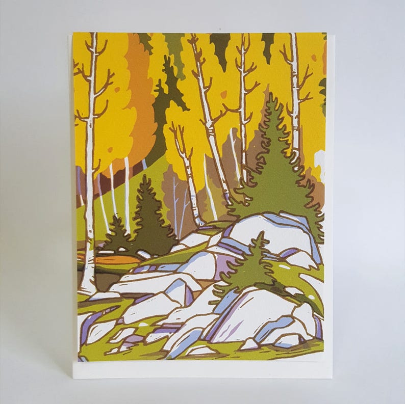 May include: A greeting card featuring a colorful forest scene. The artwork depicts trees with yellow and orange foliage, white birch trees, and large, light-colored rocks. Green pine trees and grass add contrast to the landscape.