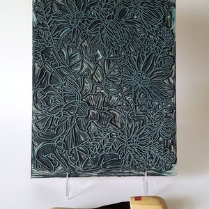 May include: A close-up of a woodblock print featuring a detailed design of stylized flowers in shades of blue and black. The print is mounted on a white surface and two wooden carving tools are in the foreground.