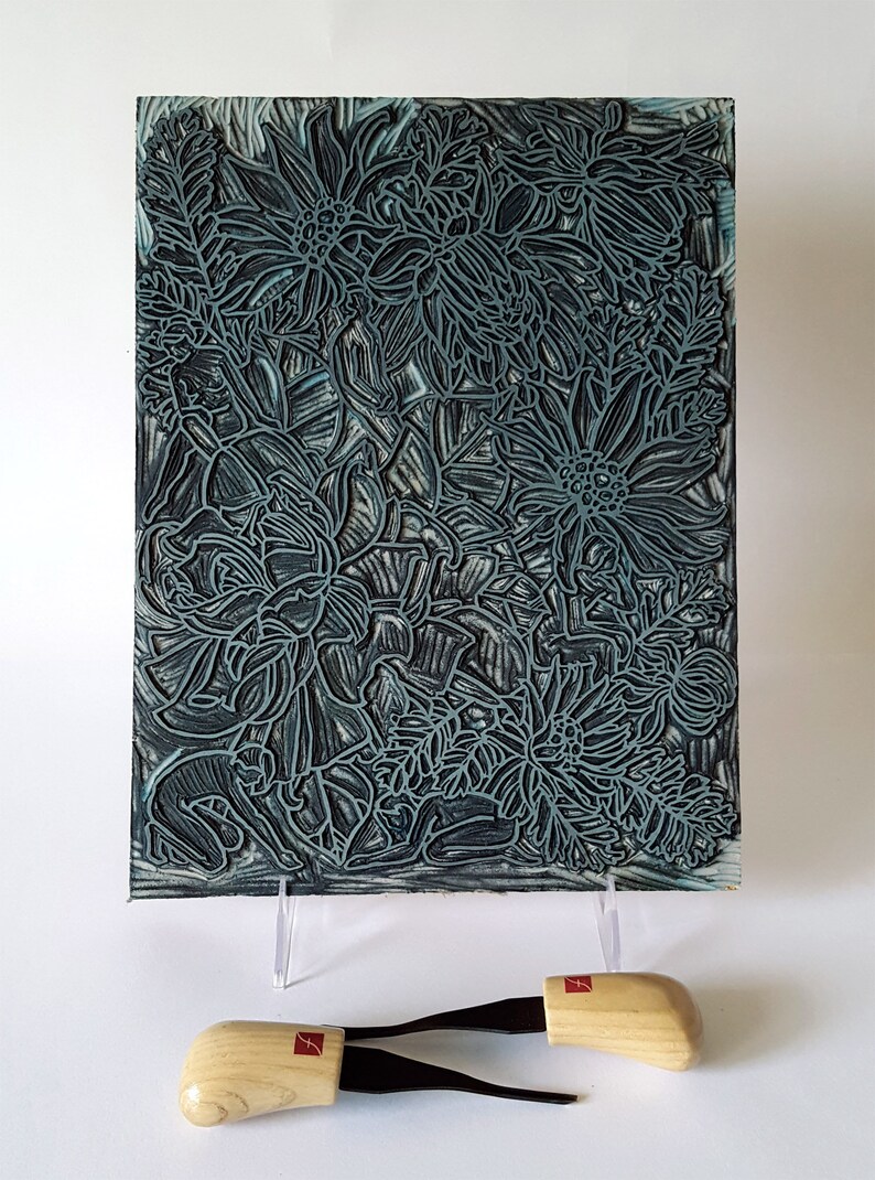 May include: A close-up of a woodblock print featuring a detailed design of stylized flowers in shades of blue and black. The print is mounted on a white surface and two wooden carving tools are in the foreground.