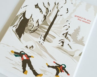 May include: A winter-themed greeting card. The illustration shows two figures in dark outfits creating snow angels. The background depicts a snowy landscape with trees, and the text "grateful for you this season" is in red.