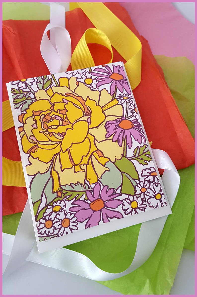 May include: A white card with a floral design featuring a large yellow flower with purple and white flowers surrounding it. The card is wrapped in white ribbon and placed on top of a stack of tissue paper in shades of green, yellow, and pink.