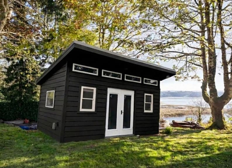 May include: A small, detached structure with dark brown horizontal wood siding, a slanted roof, and white trim. It features a set of double doors, two side windows, and a row of four rectangular transom windows positioned above the door frame.