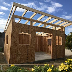 May include: A wooden shed structure under construction featuring oriented strand board walls, an open-frame roof with exposed rafters, and a large central doorway opening. The building is situated outdoors with visible window cutouts in the walls.