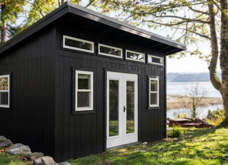 May include: A black shed with vertical siding, a slanted roof, and white-trimmed windows. The structure features a set of double doors with glass panels, four rectangular transom windows above the doors, and two side windows with white frames.