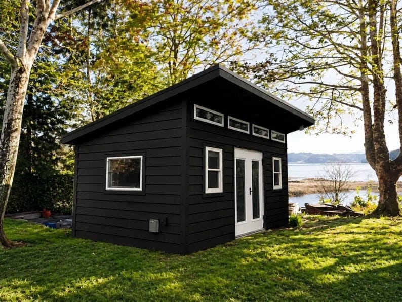 May include: A small, detached shed with black horizontal siding and a single-slope roof. The structure features white-framed windows, a set of white double doors, and a row of four small rectangular transom windows located near the roofline.