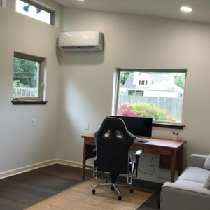 May include: A home office space featuring a wooden desk with a black computer monitor and a black ergonomic gaming chair. A light gray sofa with patterned throw pillows sits to the right. The room has white walls, dark wood floors, and windows.
