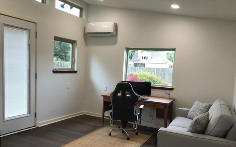 May include: A home office space featuring a wooden desk with a black computer monitor and a black ergonomic gaming chair. A light gray sofa with patterned throw pillows sits to the right. The room has white walls, dark wood floors, and windows.