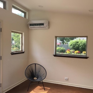 May include: A black circular chair with a woven cord seat and backrest, positioned in a room with off-white walls, a white wall-mounted air conditioning unit, a glass door, and two windows looking out onto a green lawn and garden area.