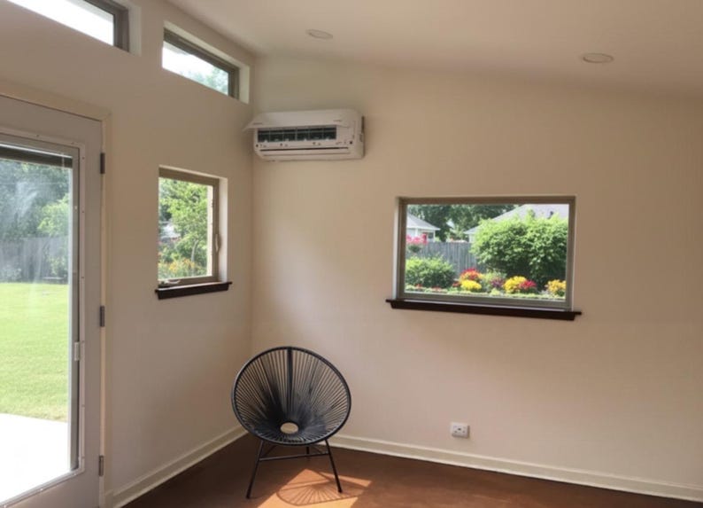 May include: A black circular chair with a woven cord seat and backrest, positioned in a room with off-white walls, a white wall-mounted air conditioning unit, a glass door, and two windows looking out onto a green lawn and garden area.