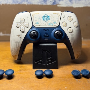 Puede incluir: Un mando personalizado de PlayStation 5 en blanco y azul marino, con detalles dorados y turquesas. El mando está sobre un soporte negro con el logotipo de PlayStation, y hay joysticks azules adicionales.