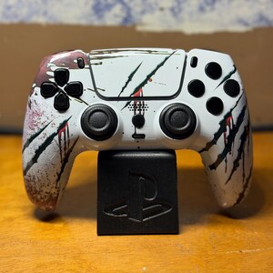 May include: A white video game controller featuring black claw mark graphics with red blood-like splatters. The controller has black analog sticks, a black directional pad, and black action buttons, resting on a black stand with a PlayStation logo.