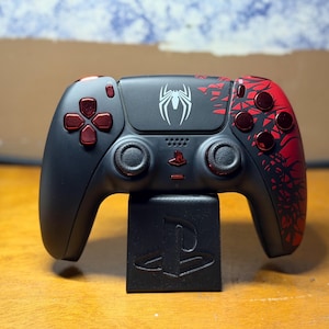 May include: A black and red video game controller featuring a white spider emblem on the central touchpad and a black stand with an embossed PlayStation logo. The controller includes metallic red directional buttons and a red web-like graphic pattern.