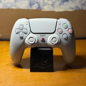 Puede incluir: Un mando de juego inalámbrico blanco con un logotipo de PlayStation central, dos joysticks analógicos, una cruceta y cuatro botones de acción con símbolos de triángulo, círculo, cruz y cuadrado, sobre un soporte negro con logotipo.