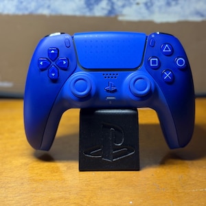 May include: A cobalt blue wireless video game controller featuring a central touchpad with a subtle geometric pattern, two analog sticks, a directional pad, and four action buttons marked with triangle, circle, square, and cross symbols.