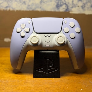 May include: A lavender and white game controller with a black stand. The controller has a symmetrical design with buttons and joysticks. The stand is a black cube with a PlayStation logo.
