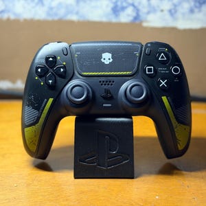 Può includere: Un controller di gioco wireless nero con un'icona a forma di teschio bianco sul touchpad centrale, strisce gialle e impugnature testurizzate. Il controller è posizionato su un supporto nero con il logo PlayStation e presenta i tasti standard.
