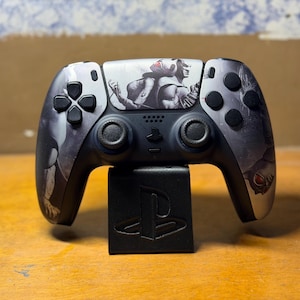 May include: A black and grey wireless video game controller featuring a graphic illustration of a character on the central touchpad. The controller is mounted on a black stand embossed with the PlayStation logo, featuring dual analog sticks and buttons.