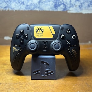 May include: Black gaming controller with yellow detailing and text, on a black stand with a logo. The controller has buttons, a directional pad, and analogue sticks. The text on the controller reads "DREABRIDGE".