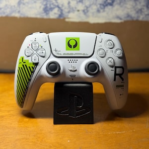 May include: A white wireless video game controller featuring a green square logo, black analog sticks, and directional buttons. The left grip has green diagonal stripes and text, while the right side displays a large black letter R and symbols.
