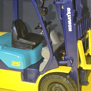 May include: A blue and yellow toy forklift with the brand name "Komatsu" printed on the side. The forklift has a black seat and a blue and yellow cab. The forklift has a 25 printed on the side.