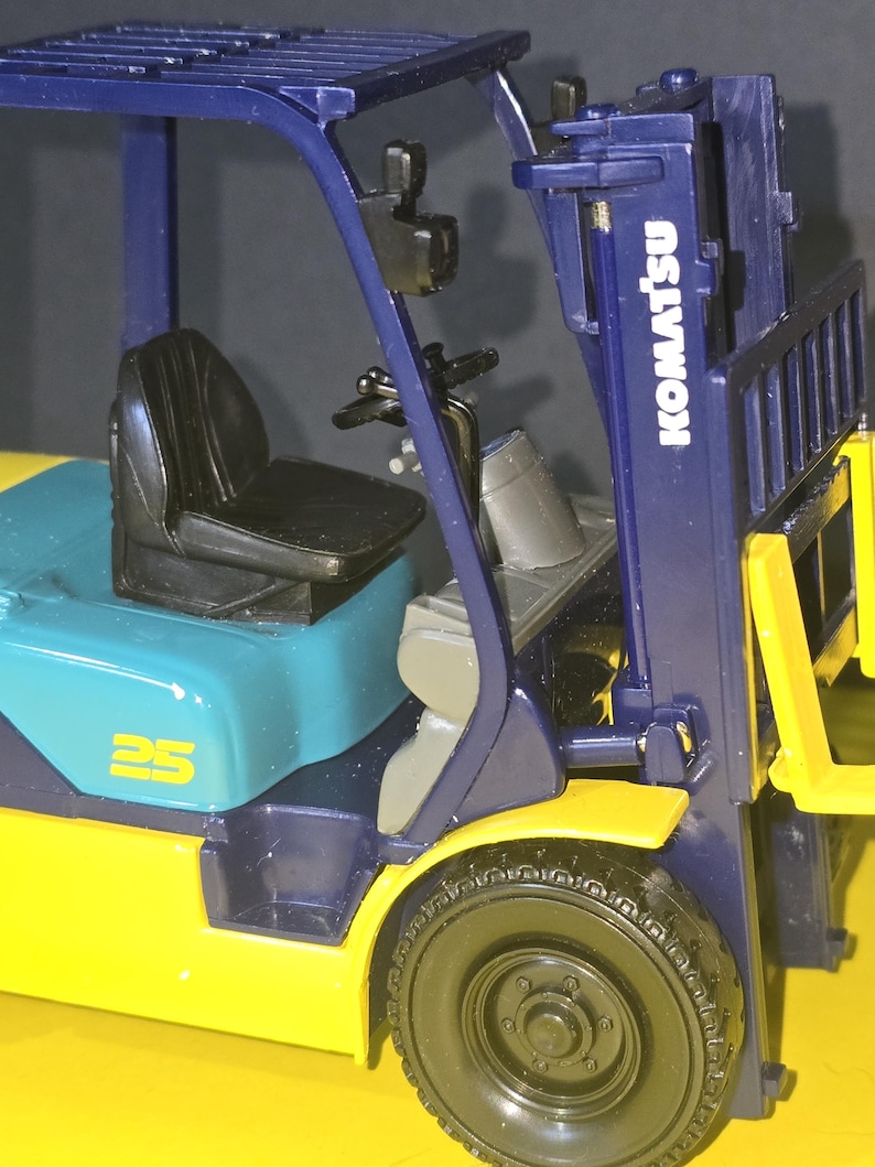 May include: A blue and yellow toy forklift with the brand name "Komatsu" printed on the side. The forklift has a black seat and a blue and yellow cab. The forklift has a 25 printed on the side.