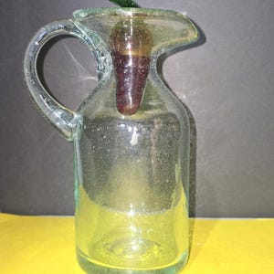 May include: A clear glass pitcher with a handle and spout. It has a dark brown stopper and a green glass leaf on top. The pitcher is on a yellow surface.