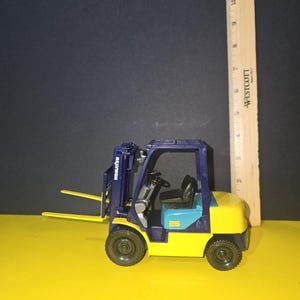 May include: A blue and yellow toy forklift with a black steering wheel and a black and white "Komatsu" logo on the side. The forklift is sitting on a yellow surface.