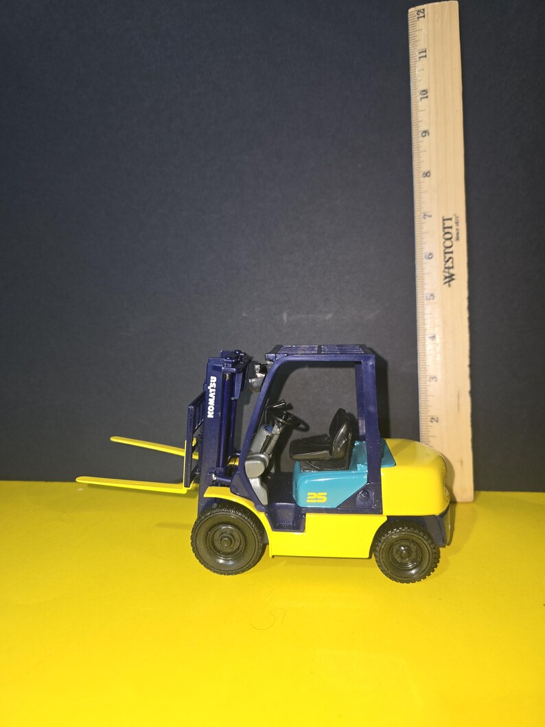 May include: A blue and yellow toy forklift with a black steering wheel and a black and white "Komatsu" logo on the side. The forklift is sitting on a yellow surface.