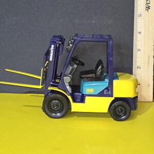 May include: A blue and yellow toy forklift with the word "KOMATSU" printed on the side. The forklift has a blue cab with a black steering wheel and a black seat. The forklift has a blue lift arm with a yellow fork. The forklift is on a yellow surface.