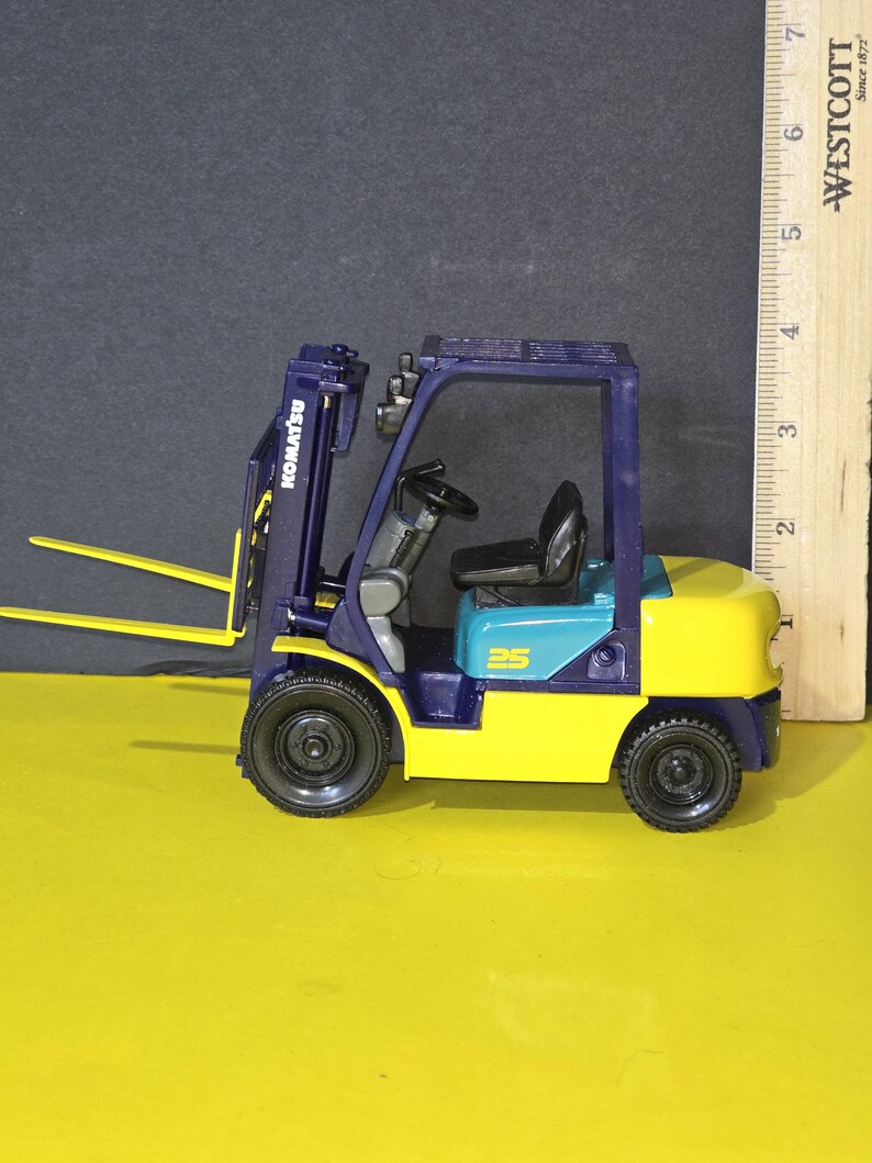 May include: A blue and yellow toy forklift with the word "KOMATSU" printed on the side. The forklift has a blue cab with a black steering wheel and a black seat. The forklift has a blue lift arm with a yellow fork. The forklift is on a yellow surface.