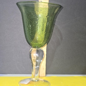 May include: A green glass goblet with a clear, bulbous stem and a wide, flared rim. The green bowl has small air bubbles. A wooden ruler is placed behind the goblet. The goblet sits on a clear, circular base.