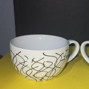 Könnte beinhalten: Weiße Keramik-Teetasse mit gebogenem Henkel. Die Tasse hat ein abstraktes, schwarzes Kritzeldesign auf der Außenseite. Die Tasse steht auf einer gelben Oberfläche.