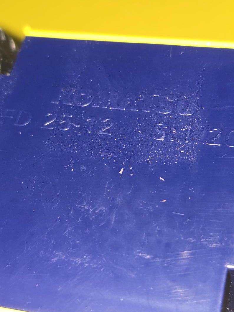May include: Close-up of a blue plastic part with the text "KOMATSU 25-12 5-1/20" embossed on it.