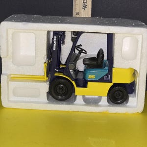 May include: A blue and yellow toy forklift with the word "KOMATSU" on the side. The forklift is sitting in a white polystyrene box.