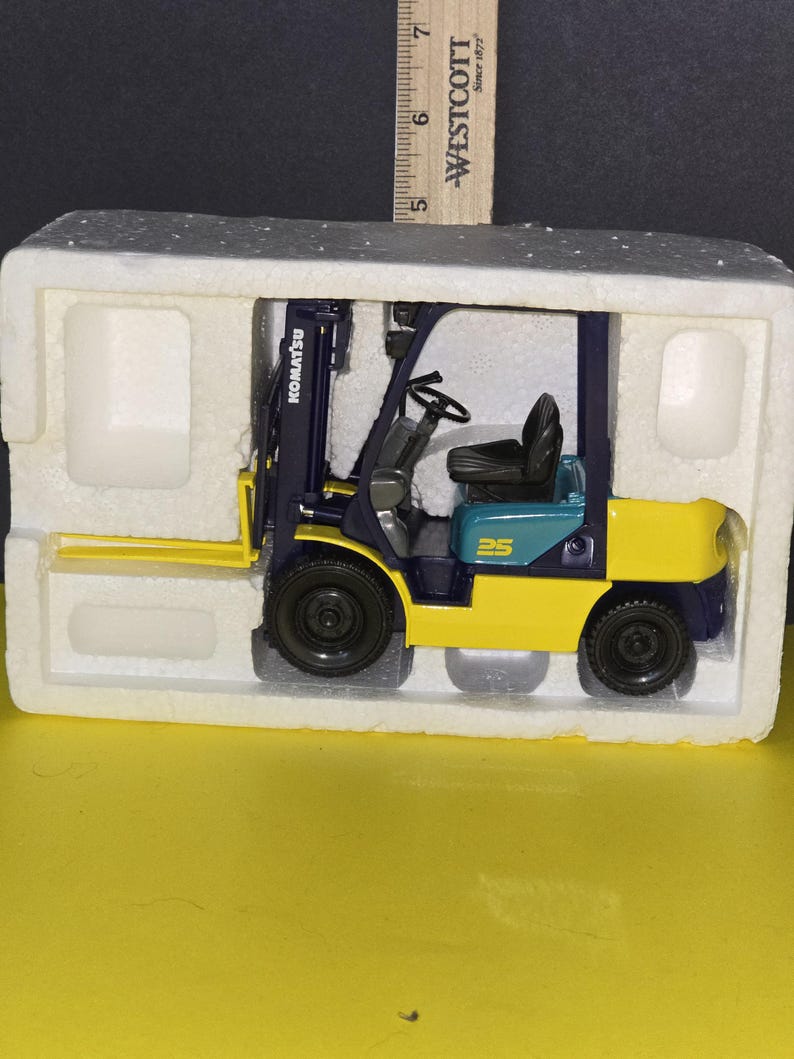 May include: A blue and yellow toy forklift with the word "KOMATSU" on the side. The forklift is sitting in a white polystyrene box.