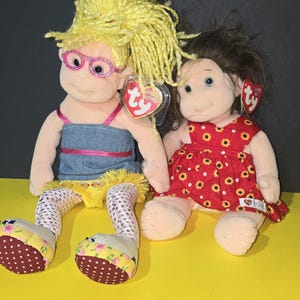 May include: Two plush dolls, one with blonde hair wearing glasses and a denim dress, and the other with brown hair wearing a red dress with yellow flowers. Both dolls have Ty tags.