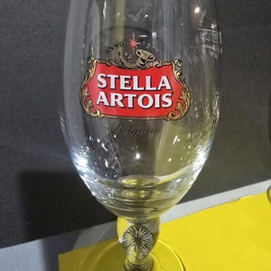 May include: A clear Stella Artois beer glass with a gold rim. The glass displays the Stella Artois logo in red and gold, with the word "Belgium" at the base. The glass has a decorative stem and a wide base.