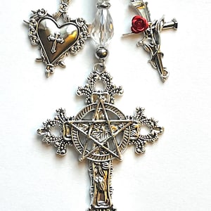 May include: A silver-toned pendant featuring a large ornate cross with a central pentagram. Above the cross hang three strands of clear faceted beads and silver spacers. The side charms include a heart with a cross and a nail with a red rose.