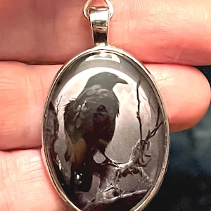 Puede incluir: Un colgante ovalado que muestra un cuervo negro posado en una rama frente a una luna llena. La imagen está enmarcada en un metal plateado pulido con un borde liso y redondeado, con una anilla en la parte superior para una cadena.