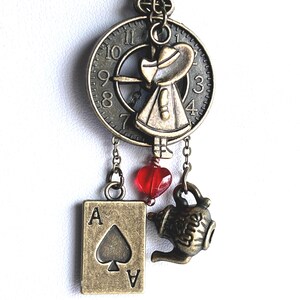 May include: A bronze-toned necklace pendant featuring a circular clock face charm with a small figure, a rectangular ace of spades playing card charm, a miniature teapot charm, and a small translucent red heart-shaped bead hanging from chains.
