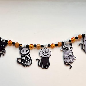 May include: A Halloween garland with cat, pumpkin, mummy, and ghost charms. The charms are black, grey, and white, strung on a beaded strand with orange and black beads. Ideal for festive holiday decorations.