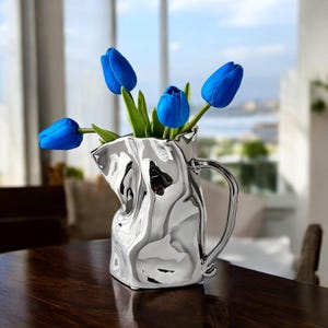May include: A silver, crumpled, ceramic vase with a handle, filled with blue tulips.