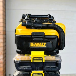 Könnte beinhalten: Ein Stapel schwarzer und gelber DEWALT ToughSystem Werkzeugkisten und ein Staubsauger. Das obere Gerät ist ein gelb-schwarzer Staubsauger mit einem schwarzen Schlauch. Die mittleren und unteren Geräte sind schwarze Werkzeugkisten mit gelben Verschlüssen und dem ToughSystem-Logo.