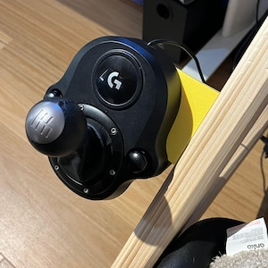 Op de afbeelding: Zwarte en zilveren versnellingspook met een zwarte basis, gemonteerd op een gele ondergrond. De versnellingspook heeft een zilveren knop en een zwarte lederachtige hoes. Het Logitech-logo is zichtbaar aan de bovenkant.