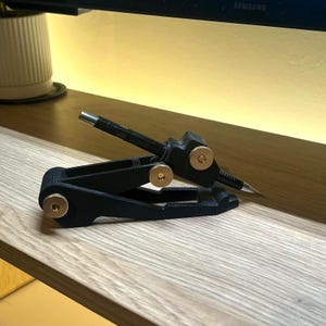 May include: A black and gold compass tool with a silver tip, designed for precise measurements. The tool features a hinged design with gold-colored adjustment knobs. It is resting on a light-colored wooden surface.