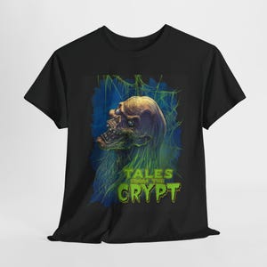 Puede incluir: Camiseta negra con un gráfico de "Tales from the Crypt". El diseño presenta una calavera en descomposición con detalles verdes y azules, sobre un fondo oscuro. Las palabras "TALES FROM THE CRYPT" son verdes.