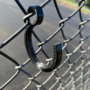 Peut inclure: Un crochet en plastique noir est fixé à une clôture en treillis métallique. Le crochet est en forme de lettre 'C' avec une petite boucle en haut.