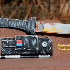 Könnte beinhalten: Ein schwarzes und silbernes Requisitenmesser mit einem gelben und orangefarbenen Griff und einer schwarzen und silbernen Scheide. Die Scheide hat einen roten, blauen und weißen Knopf an der Seite. Das Messer ist mit "Faithwalk Creations" beschriftet.