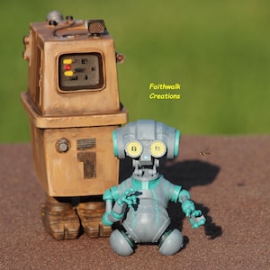 May include: Two robot figurines, one tall and boxy in tan, the other smaller and gray with teal accents. The larger robot has a screen with yellow and red lights. The smaller robot has large yellow eyes. The text "Faithwalk Creations" is visible.